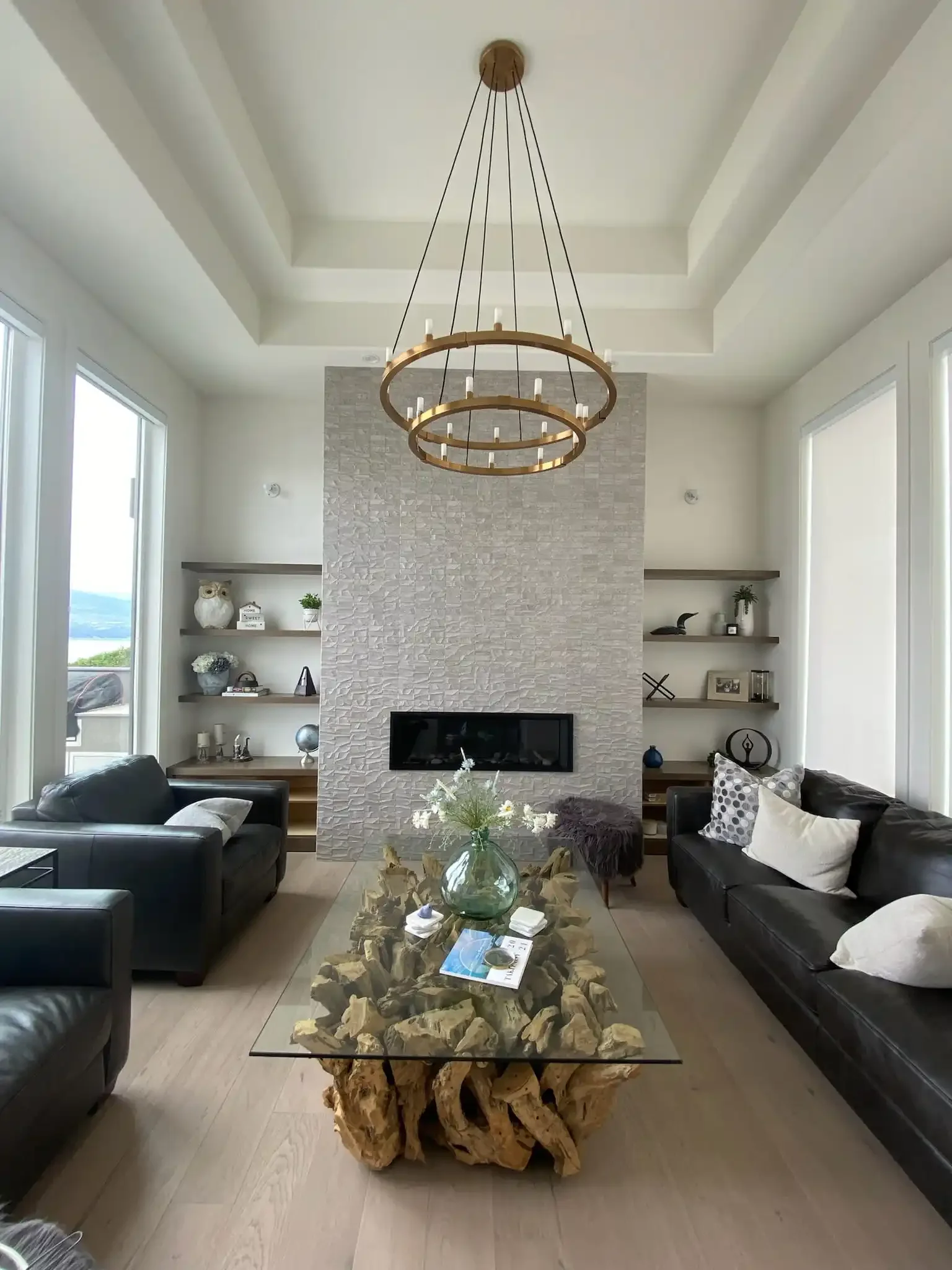 Modern living room with a white textured fireplace wall, black sofas, a glass coffee table with a green vase of white flowers, and built-in shelves on both sides of the fireplace with decorative items. A large gold circular chandelier hangs from the ceiling.