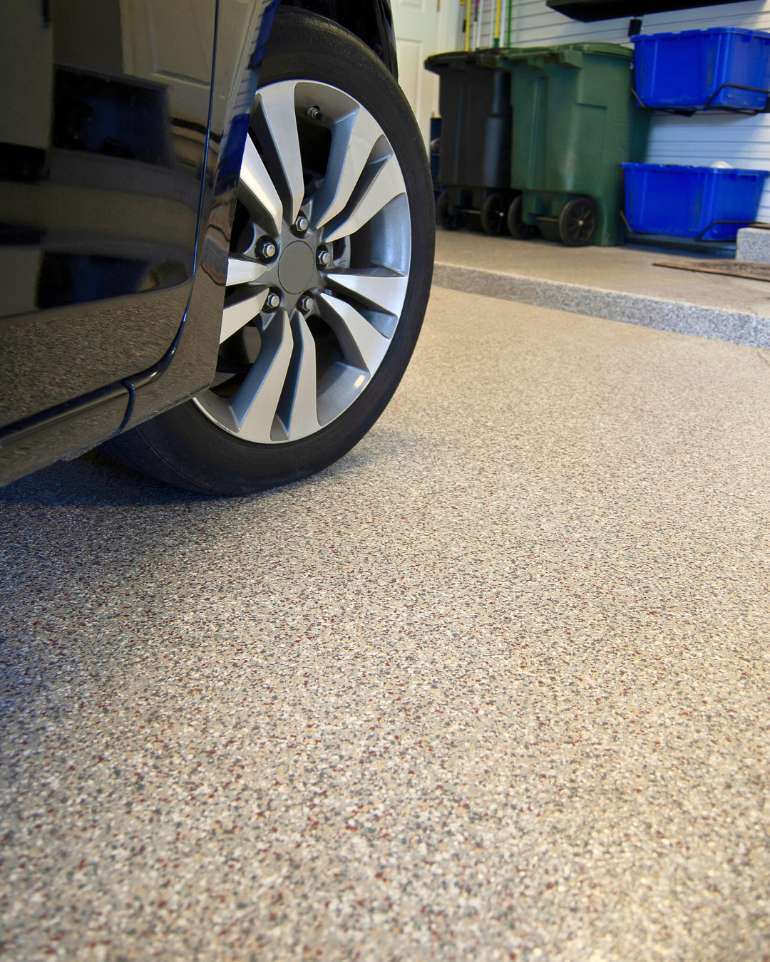 Close-up of a car tire and alloy wheel parked on a textured garage floor with storage bins in the background.
