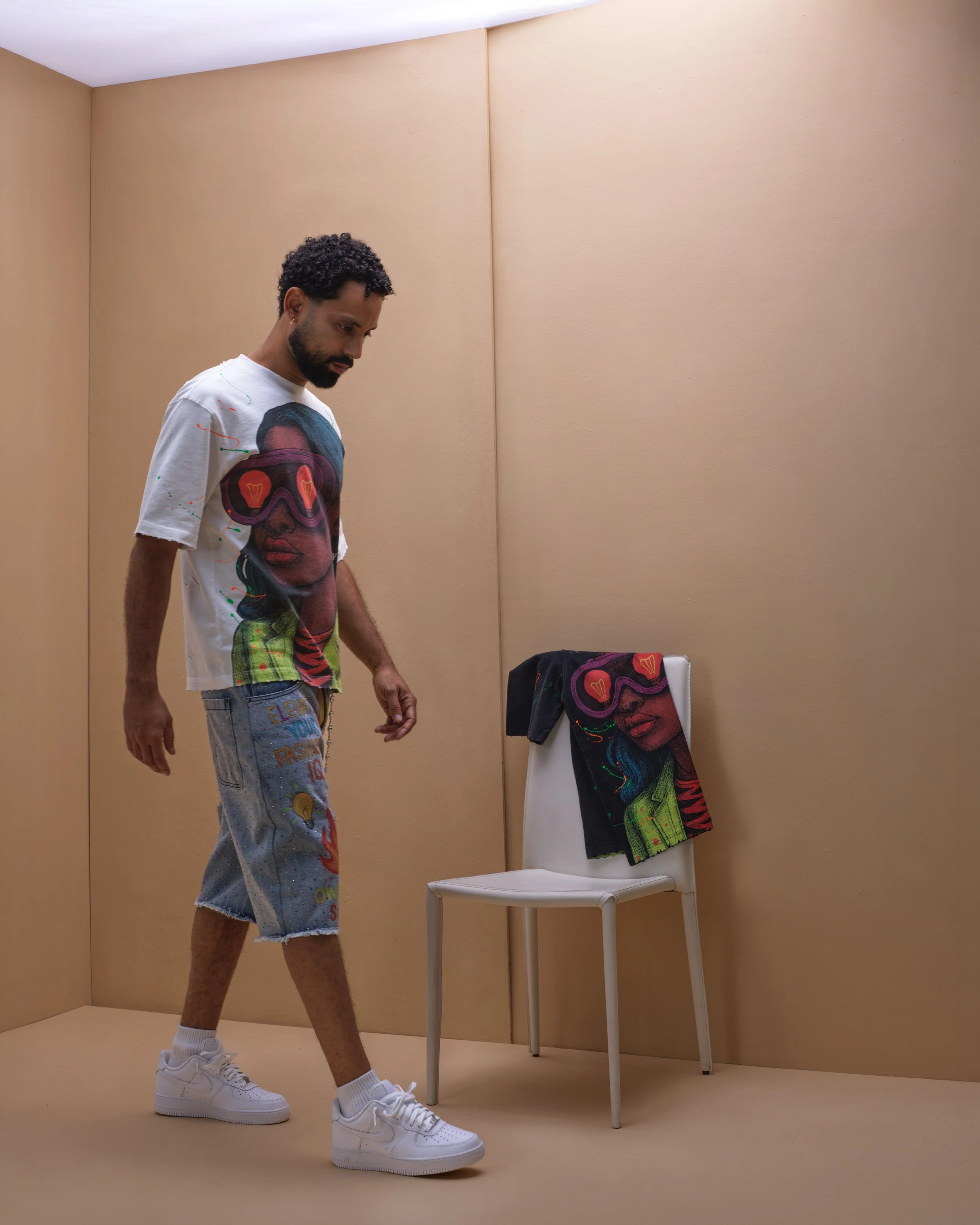 A man with curly hair and a beard looking down in a minimalist room with beige walls. He's wearing a colorful graphic t-shirt, denim shorts, white sneakers, and white socks. A chair with a matching graphic t-shirt and a black cloth draped over its ba