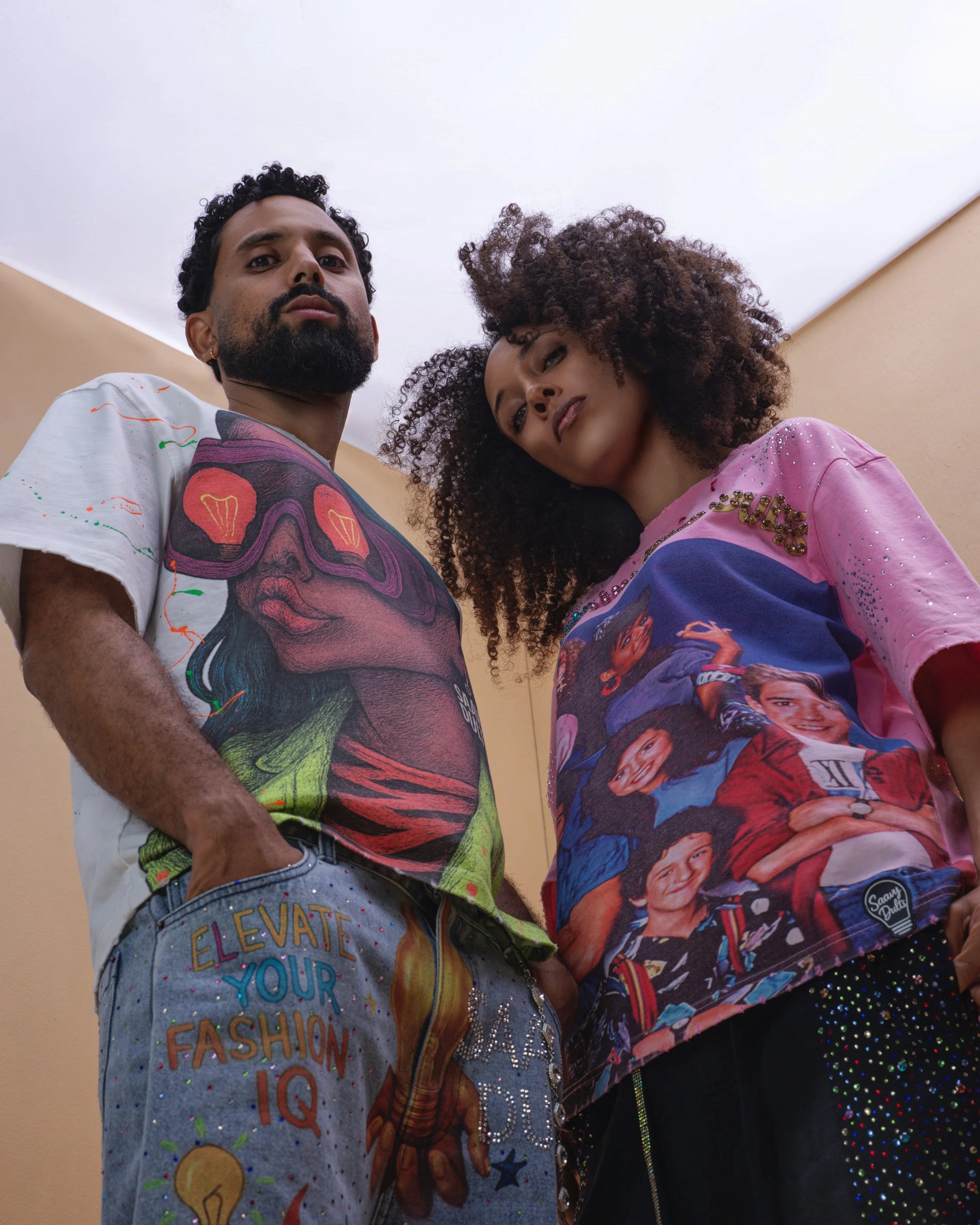 Two young adults looking down at the camera, wearing colorful graphic T-shirts with artistic and playful designs.