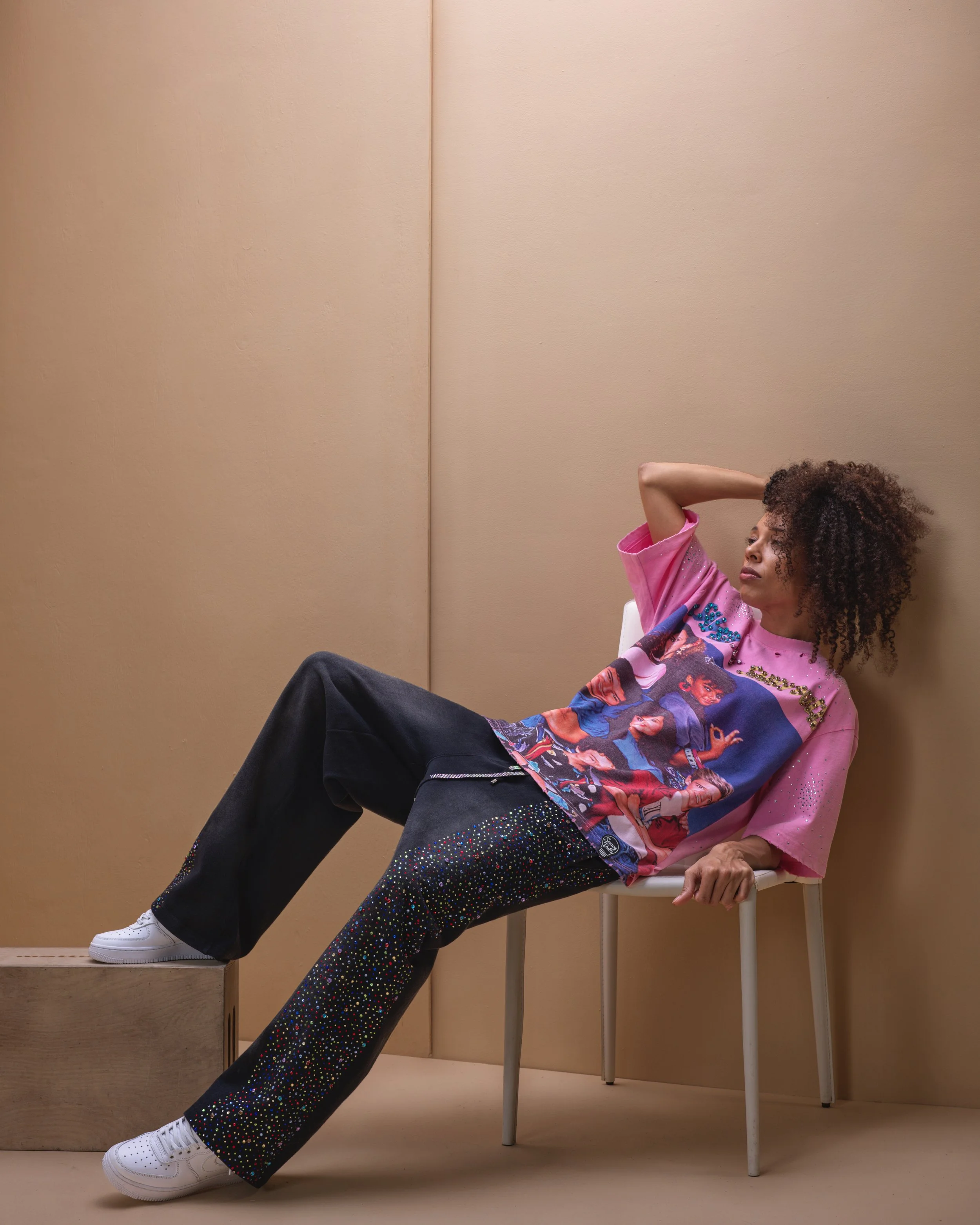A woman sitting on a chair with her head tilted back, one arm behind her head, wearing a colorful graphic T-shirt, wide-leg black pants with multicolored polka dots, and white sneakers, in a minimalist beige room.