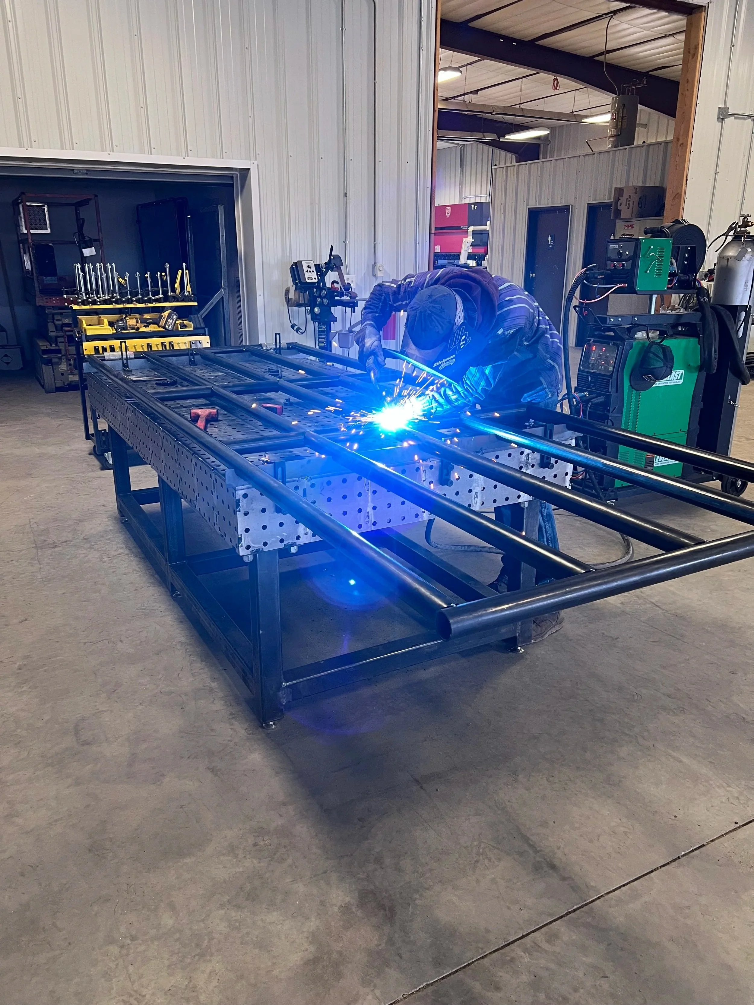 A person welding metal pipes in a workshop, creating sparks, with tools and welding equipment in the background.