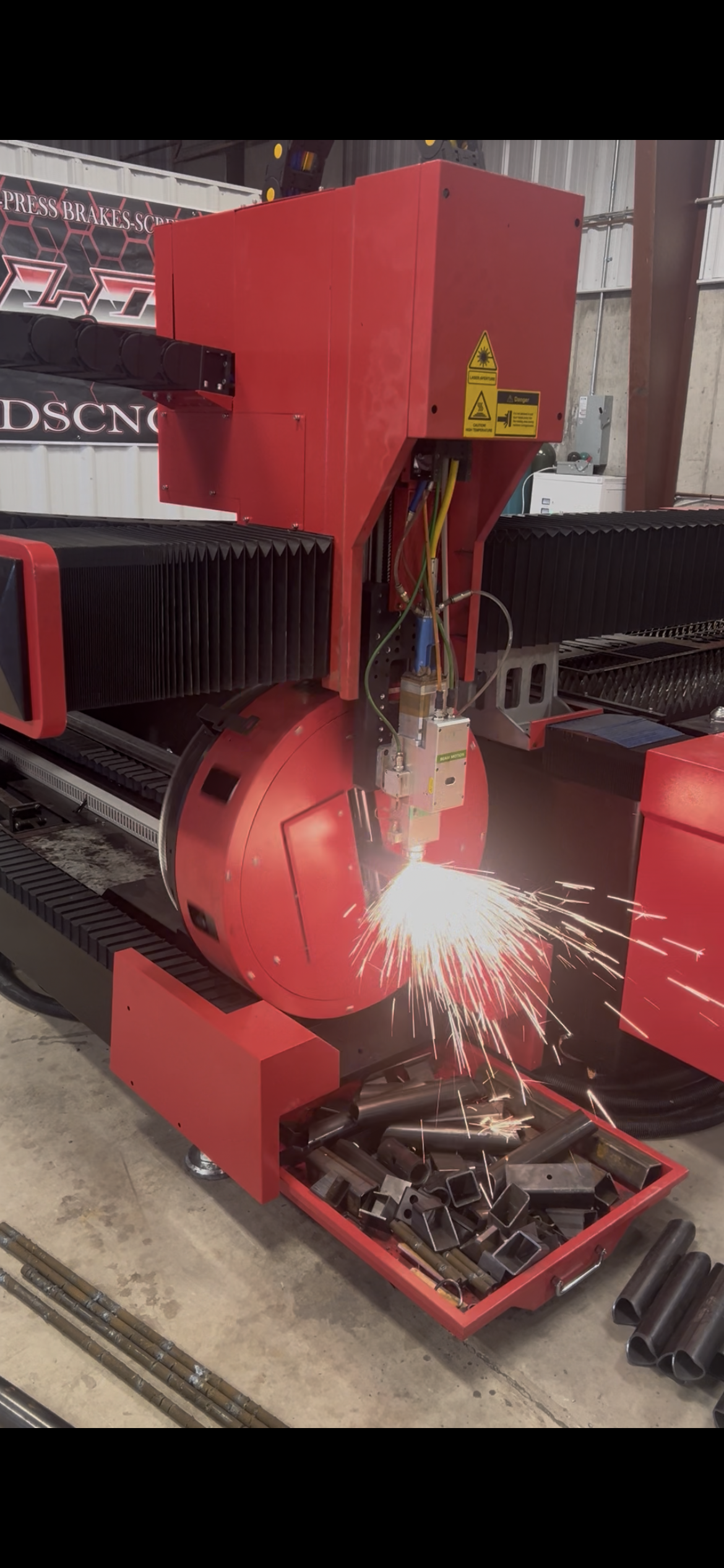 A red CNC laser cutting machine in a workshop cutting metal, with sparks flying from the cutting point, and storage box of metal parts below.