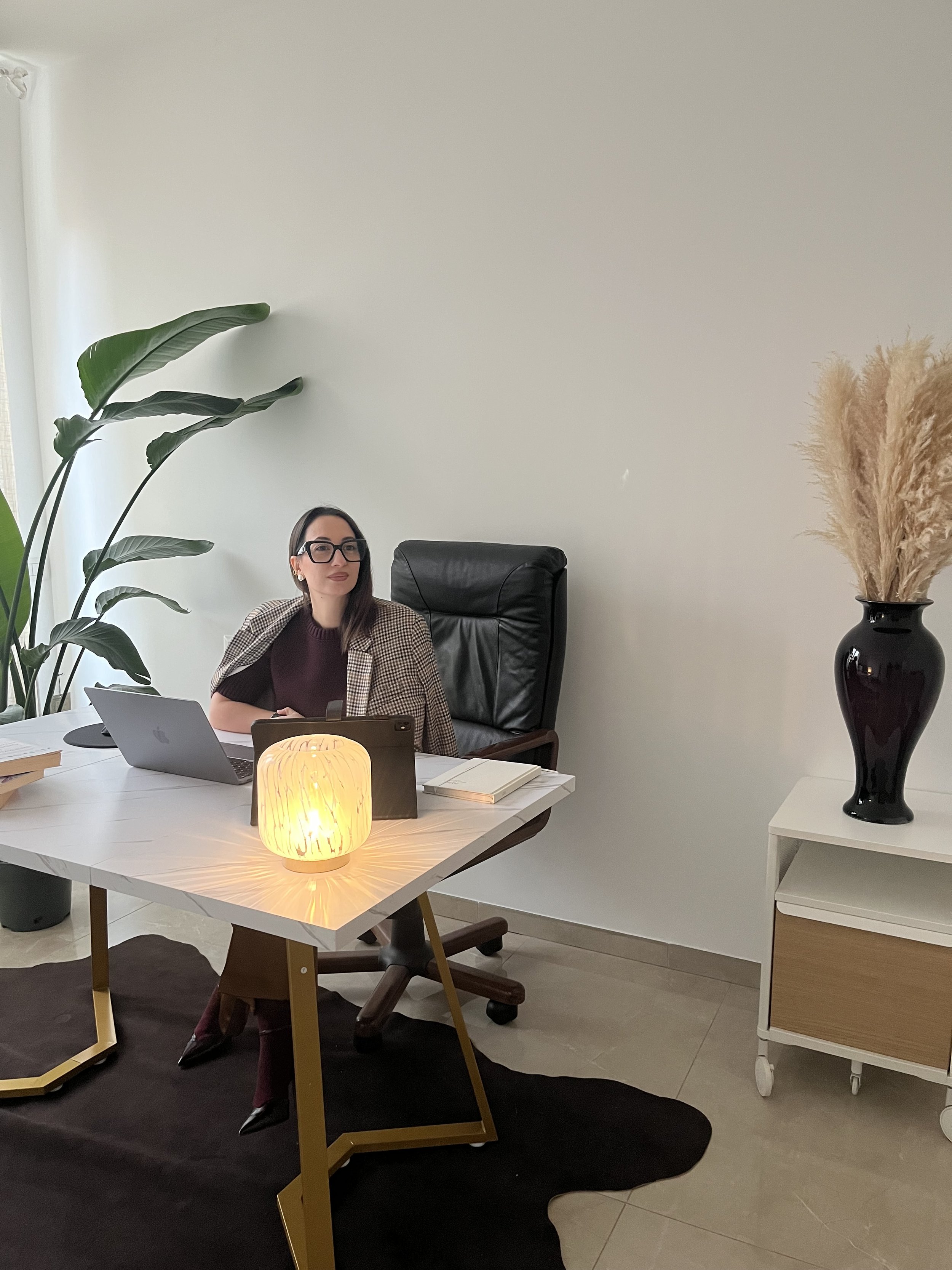 Una donna seduta dietro una scrivania in un ufficio, con una pianta grande a sinistra, una lampada accesa sulla scrivania, e un grande vassoio di libri alla sua destra. Una grande pianta e un vaso con fiori di graminacea decorano lo sfondo.