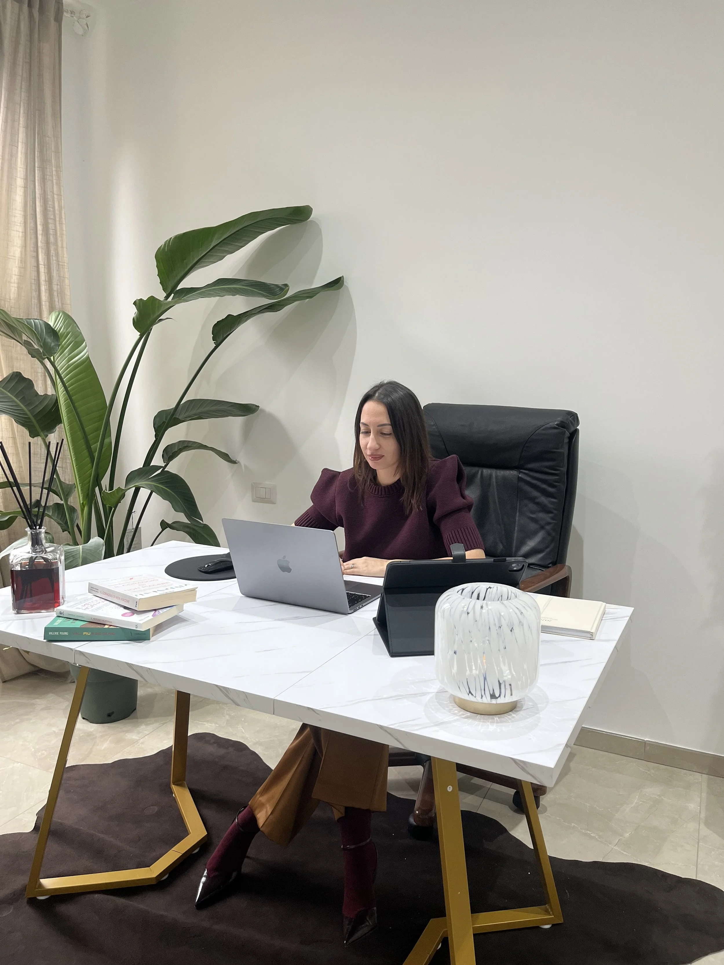 Una donna seduta alla scrivania in ufficio, con un laptop, libri, una lampada decorativa e altri oggetti, davanti a una grande pianta verde in una stanza moderna.