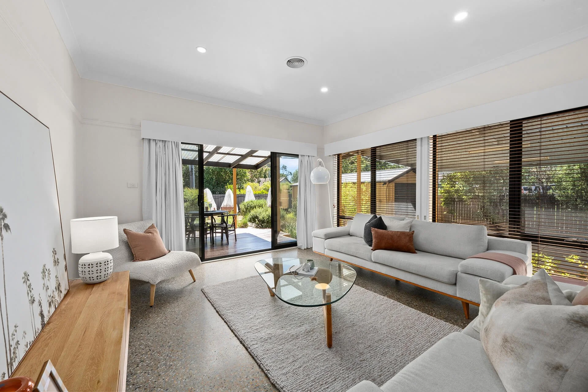 Bright living room with white sofa, armchair, glass coffee table, wooden sideboard, and large windows with blinds and curtains, opening to a garden patio with outdoor furniture.