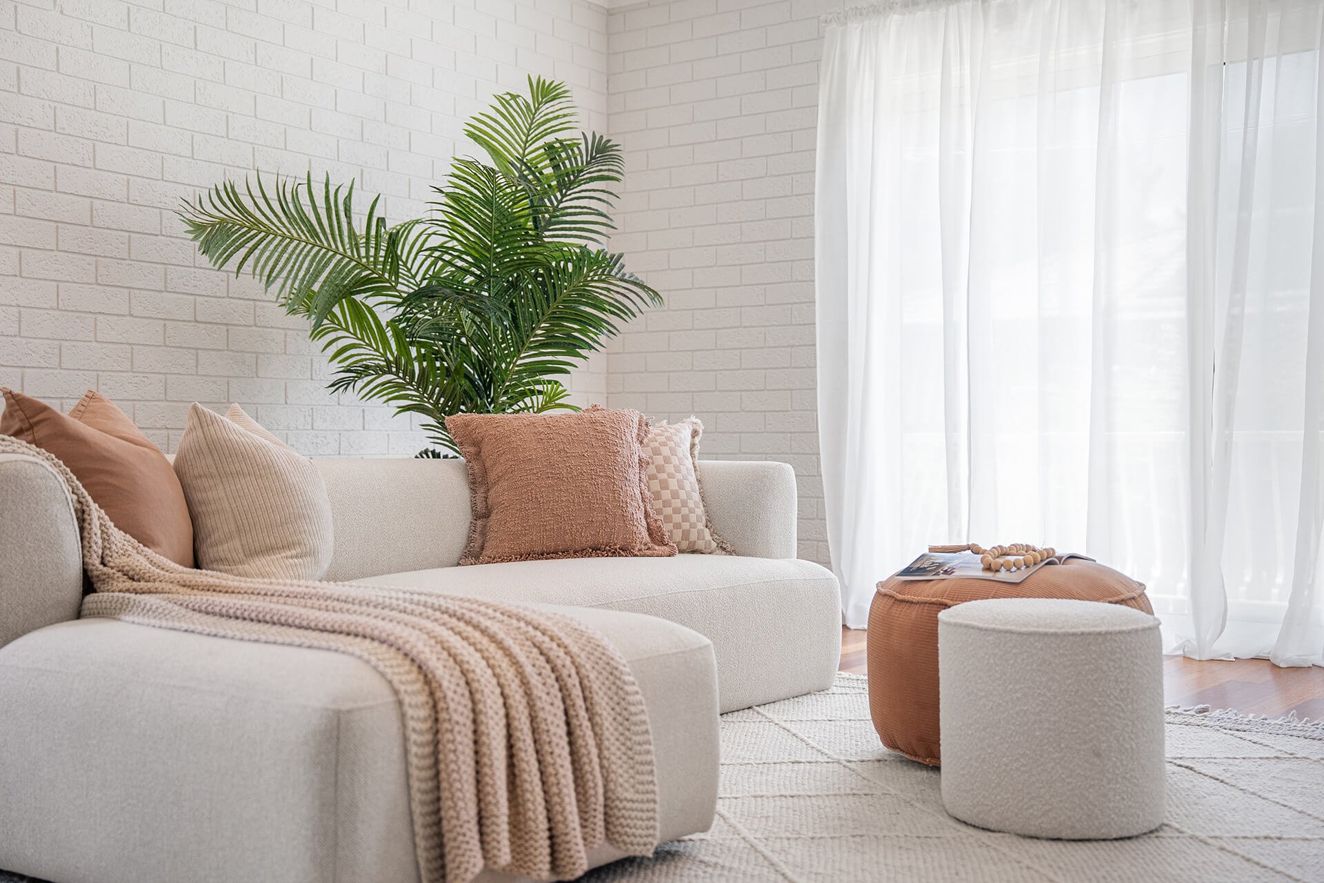 Cozy living room with a white sofa, multiple soft-colored pillows, a pink throw blanket, a large green potted plant, a round ottoman, and white curtains letting in natural light.