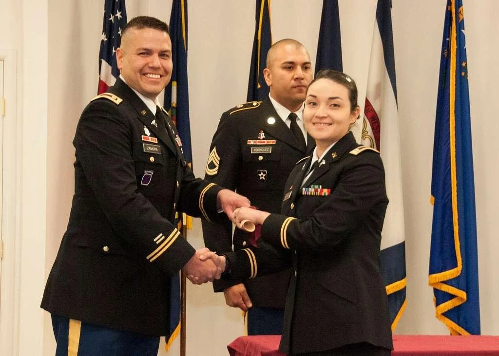 A woman in military uniform receiving a certificate or award from a smiling man in military uniform, with another man in uniform standing in the background, all in front of military flags.