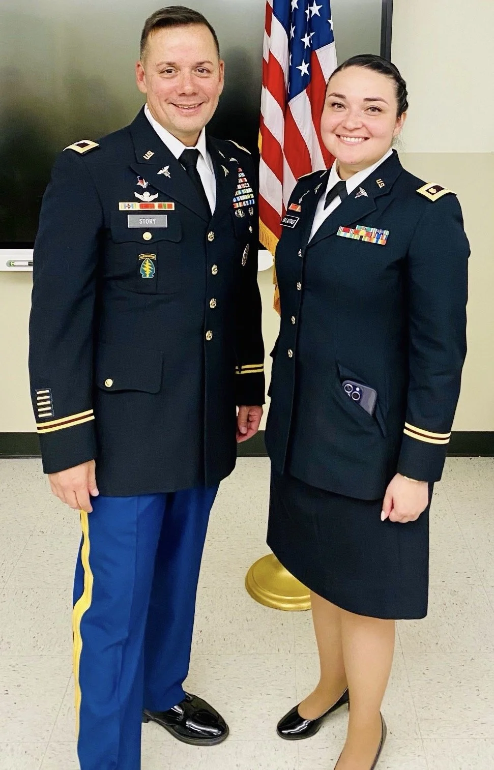 Two military officers, a man and a woman, standing indoors in front of a flag, smiling at the camera, dressed in formal military uniforms with medals and insignias.