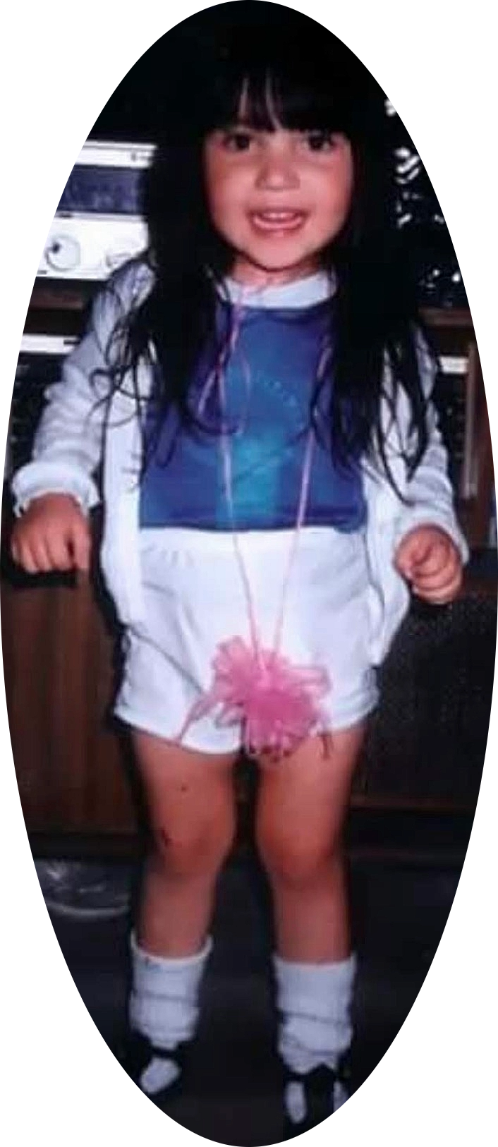 A young girl with long dark hair, wearing a white jacket, a blue shirt, white shorts, and white socks, standing indoors in front of an electronic device.