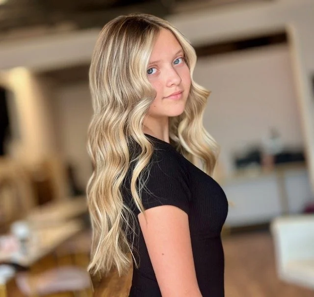 Young woman with long, wavy blonde hair wearing a black top, standing indoors with a blurred background.