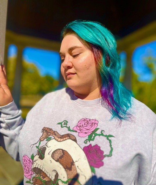 Young woman with multi-colored hair in blue, purple, and pink shades, wearing a sweatshirt with a skull and rose design, standing outdoors with a blurred background of trees and blue sky.