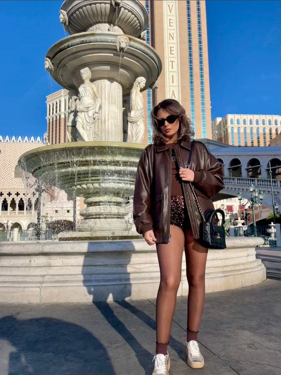 A woman in sunglasses, leather jacket, shorts, and sneakers standing in front of a decorative fountain with sculptures and water, with a tall hotel building behind her under a clear blue sky.