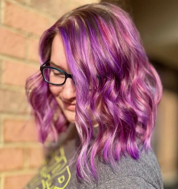 A woman with shoulder-length, wavy purple and pink hair, glasses, and a gray hoodie, smiling and standing next to a brick wall.