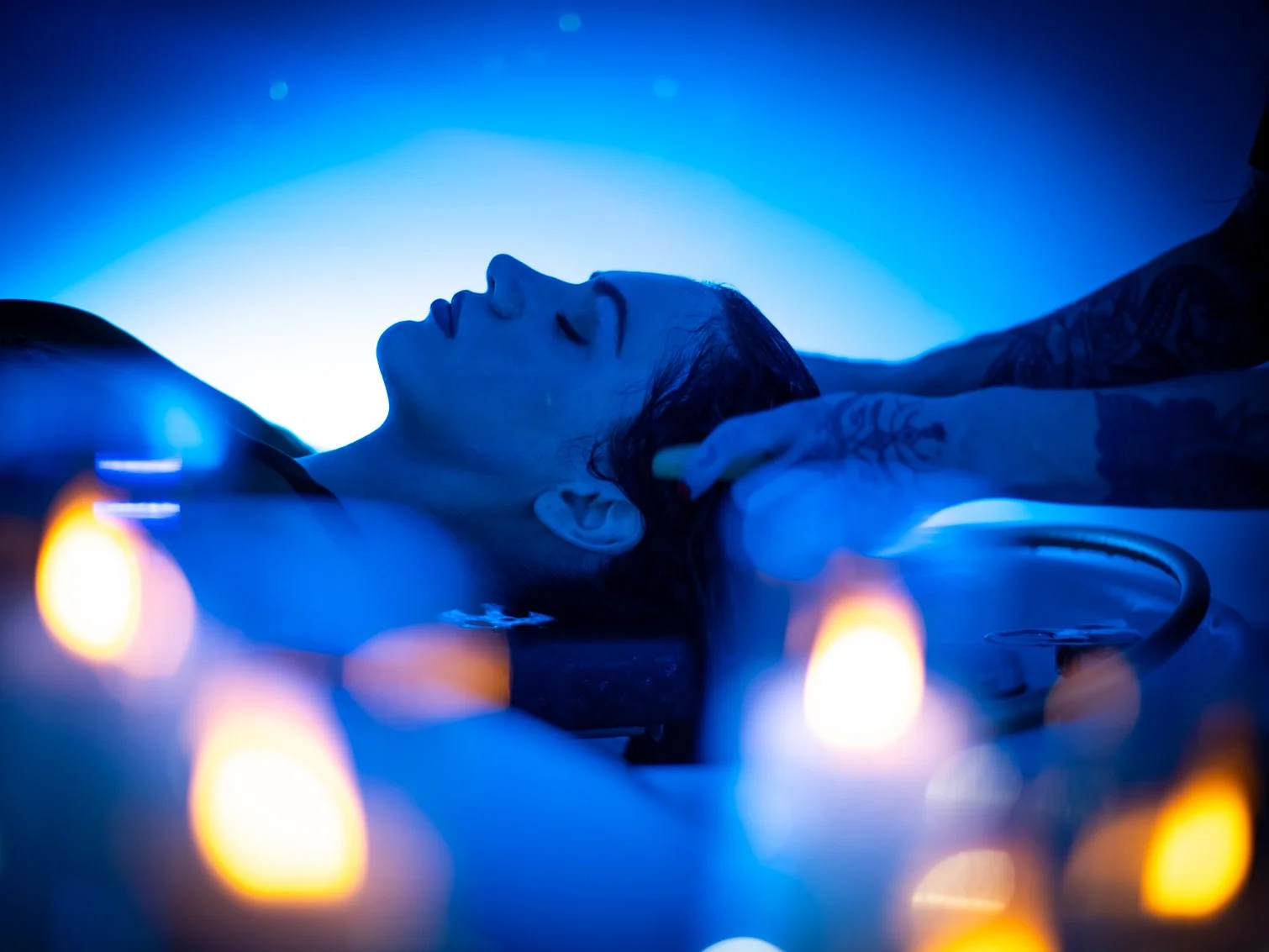 A person with closed eyes lying on their back during Japanese Head Spa treatment, being massaged or prepared for a procedure, with candles and blue lighting creating a calming atmosphere.