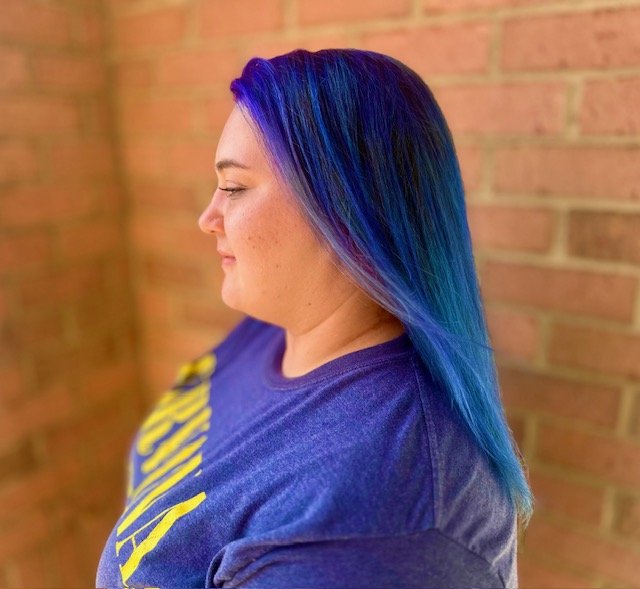 Side view of a woman with vibrant blue and purple hair, wearing a purple shirt with yellow text, standing in front of a brick wall.