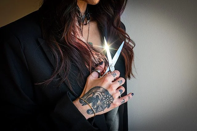 Hannah Smothers (Dyedarlin) holding a pair of scissors, with light reflecting off the blades, in front of a plain wall.