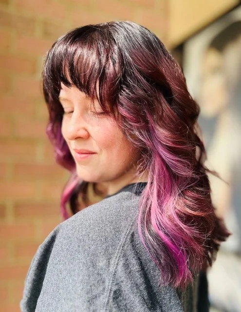 A woman with shoulder-length, dark brown hair with pink and purple highlights, smiling with her eyes closed, wearing a gray top, standing against a brick wall.