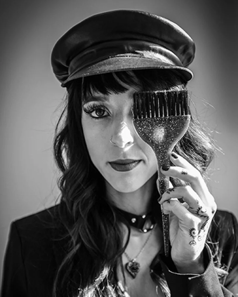 Hannah Smothers (Dyedarlin) wearing a leather cap and dark clothing, holds a hairbrush in front of her eye.