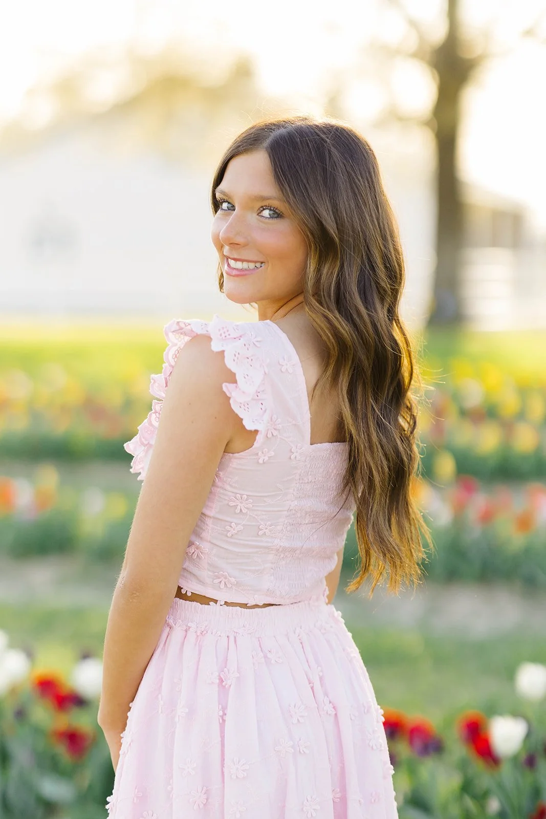 A smiling woman with long brown hair wearing a light pink dress with floral embroidery, standing outdoors in a garden with colorful flowers, during golden hour sunlight.