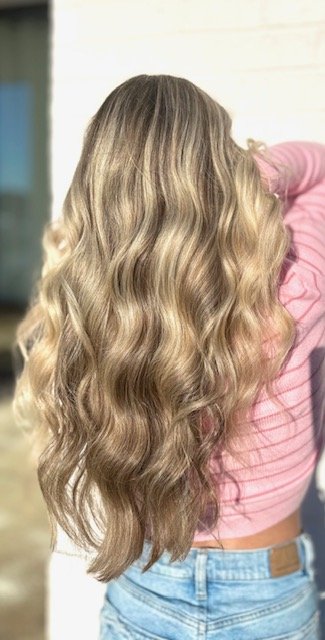 Back of a woman with long, wavy blonde hair wearing a pink top and blue jeans.