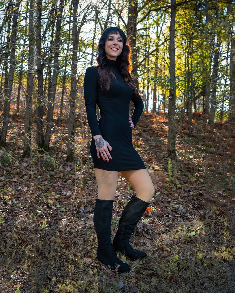 Hannah Smothers (Dyedarlin) with long dark hair wearing a black long-sleeve dress and black knee-high boots standing in a wooded area.