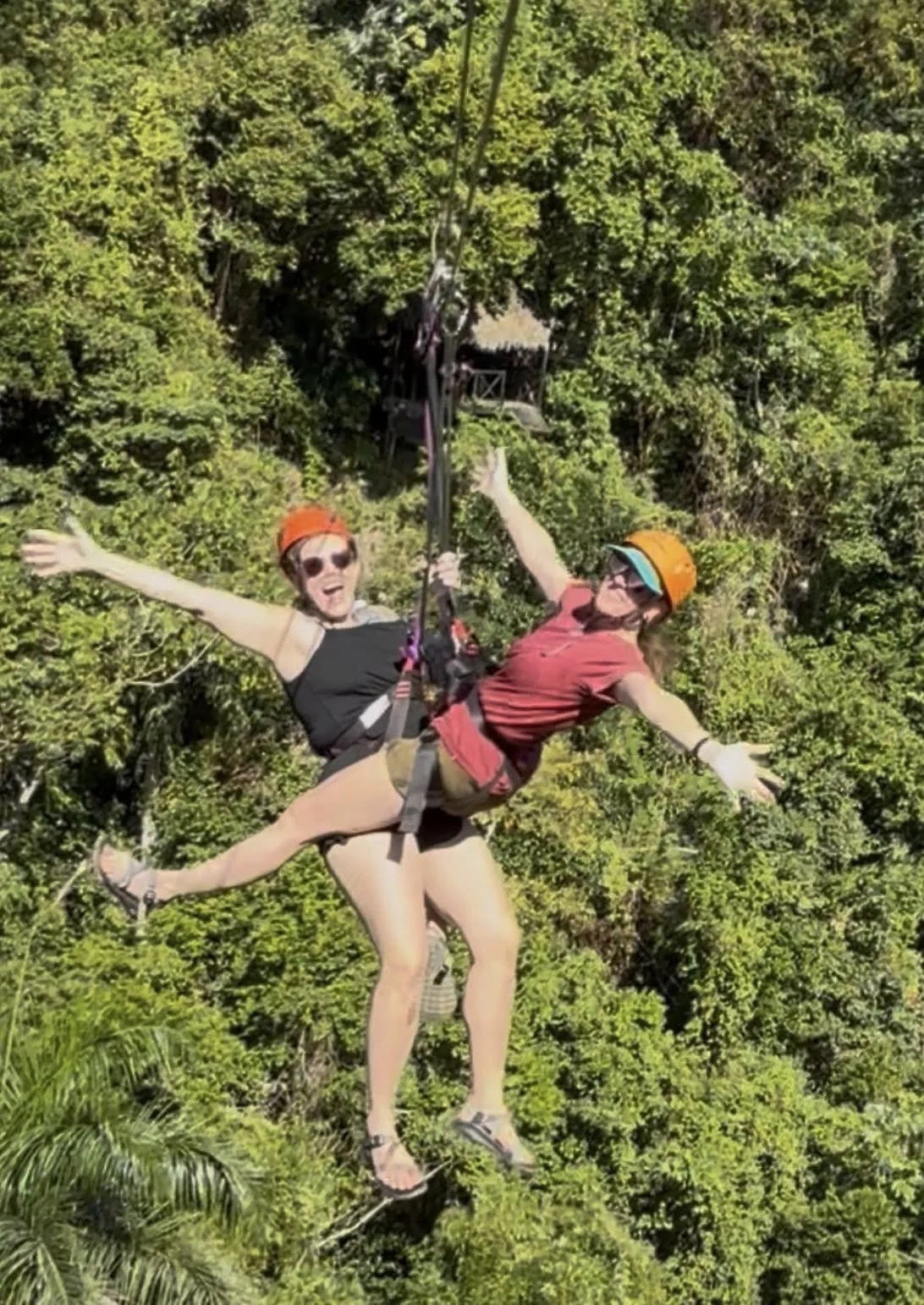 Two women are zip-lining through a lush green forest, smiling and enjoying the experience. They are wearing helmets and harnesses for safety.