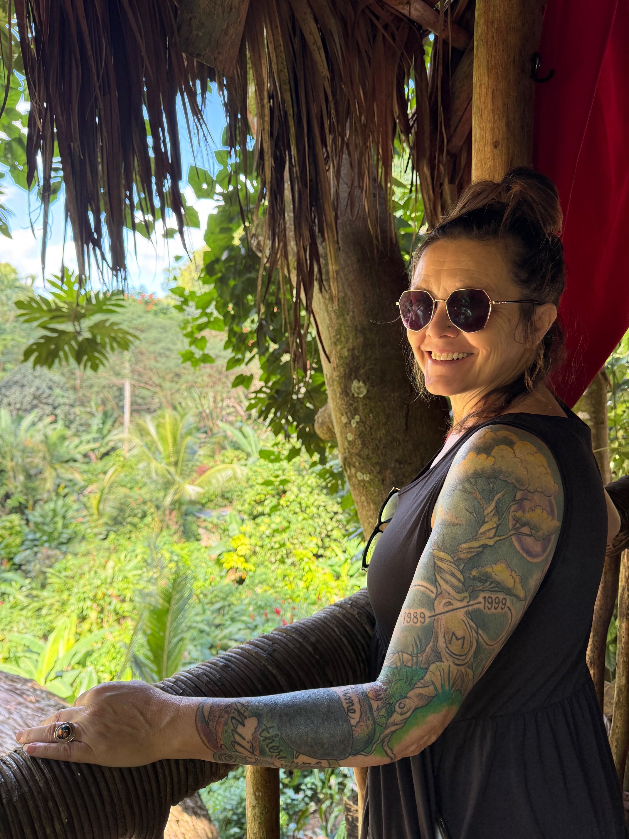 A woman with tattoos on her arms and long earrings smiling while leaning on a wooden railing in a tropical garden with lush green plants and trees, wearing sunglasses and a black sleeveless top.