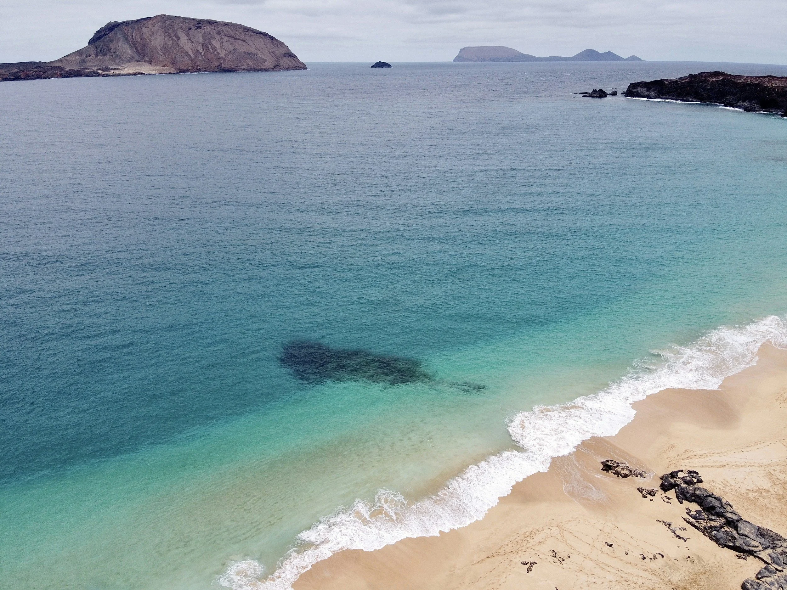 A scenic view of a beach with clear turquoise water and white sand, bordered by rocky formations, with distant islands and mountains across the horizon under a cloudy sky.