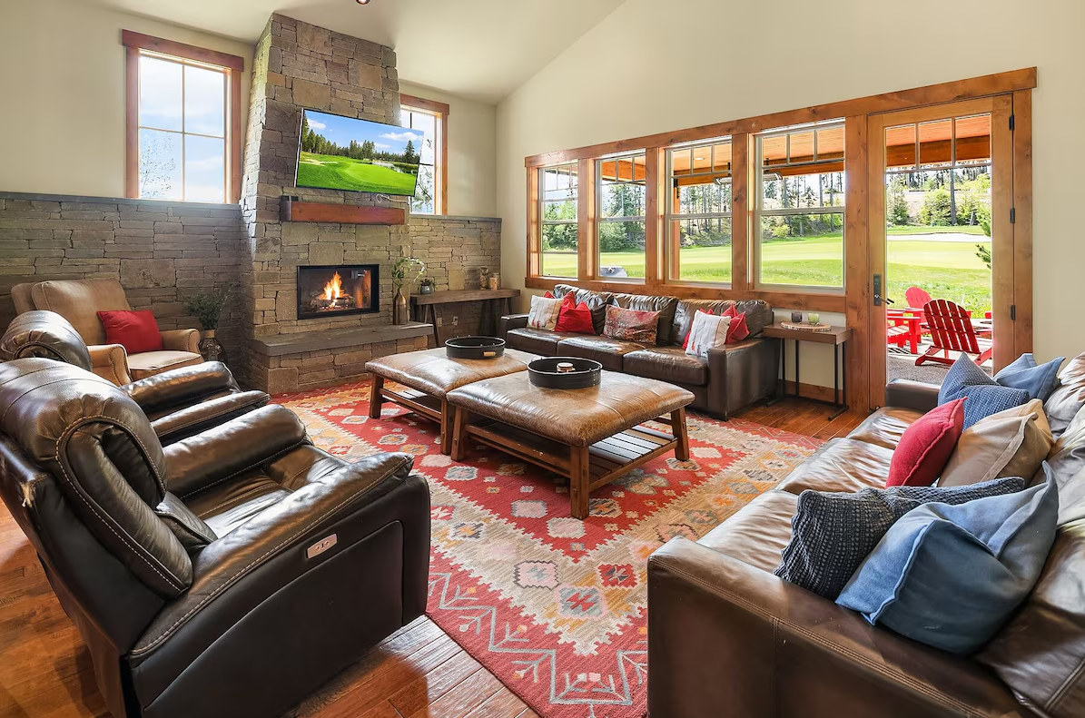 Living room with leather sofas, a stone fireplace, a TV above the fireplace, large windows, and a view of a lush green outdoor area with red chairs.