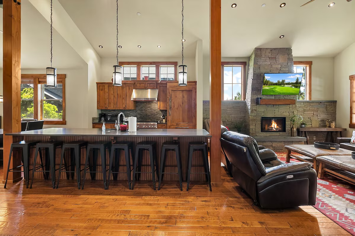 Open-concept kitchen and living room with wooden floors, wooden cabinets, a stone fireplace, and a wall-mounted TV, with black barstools at the kitchen island and leather sofas in the living area.