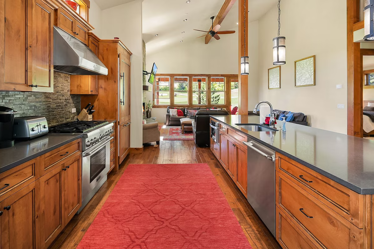 Open kitchen with wooden cabinets, black countertops, stainless steel appliances, and a red rug on wooden floor leading to living room with large windows and seating area.