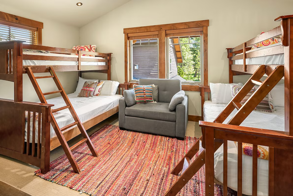 A cozy room with two wooden bunk beds, a gray armchair in the center, a colorful woven rug, and windows with wooden frames showing greenery outside.