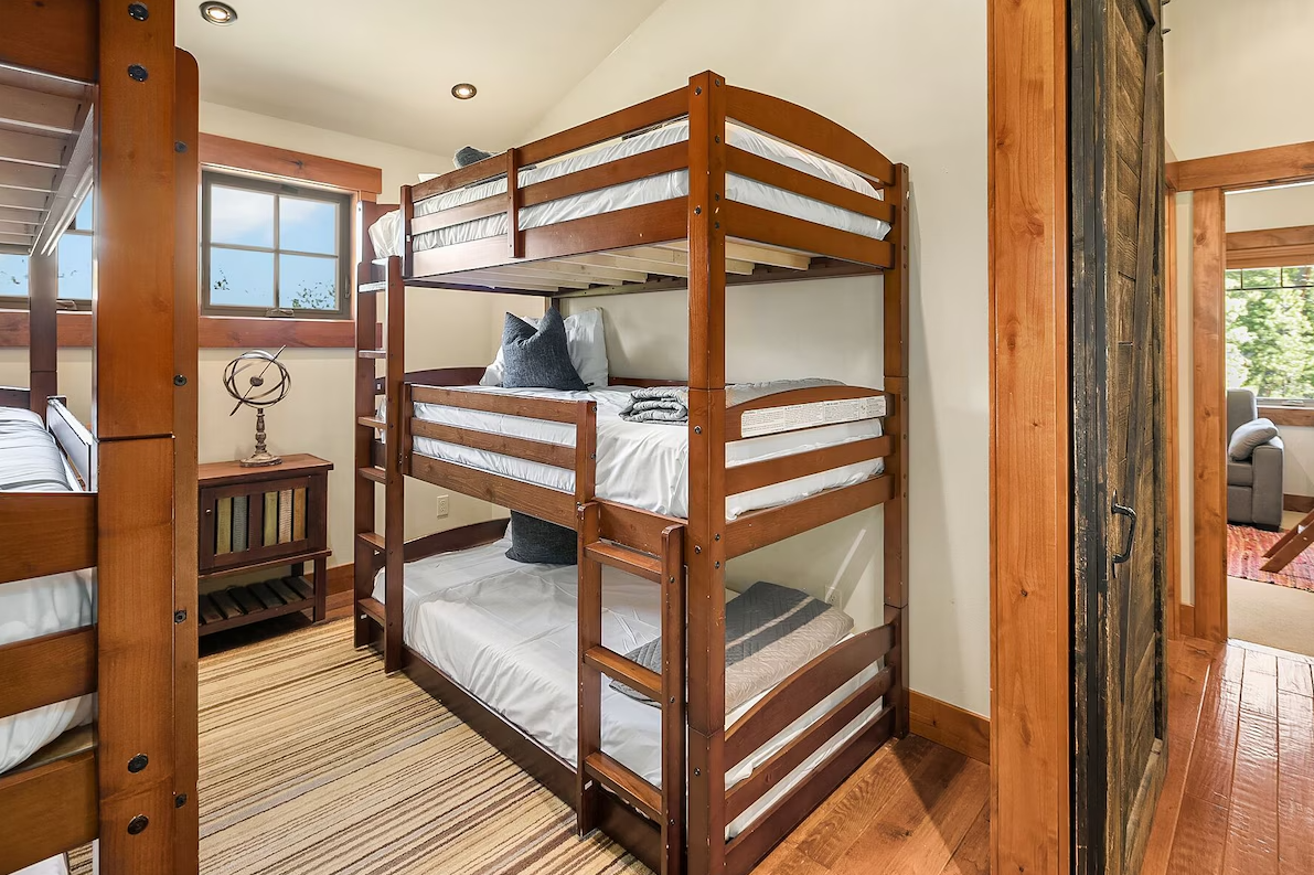 A bedroom with wood furniture, featuring a set of wooden bunk beds, a window, a small wooden nightstand with a lamp, and part of another bunk bed visible on the left. The room has wooden flooring and cream-colored walls.