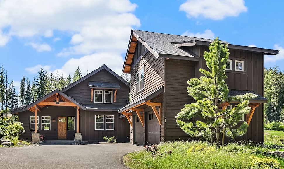 Two modern brown houses with wooden accents in a lush green area, with a clear blue sky.