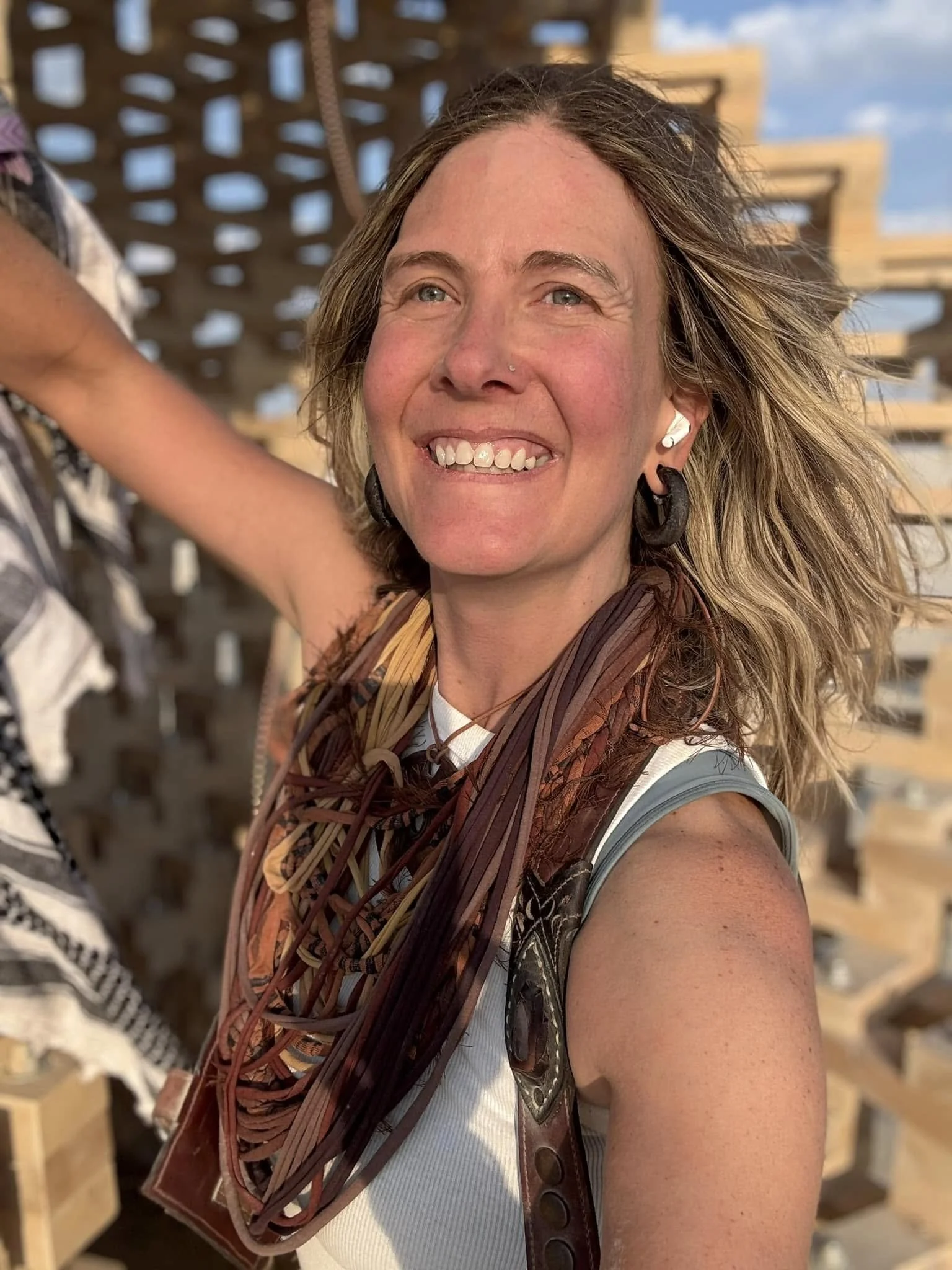 A woman smiling outdoors with a wooden structure in the background, wearing earrings, a patterned scarf, and a sleeveless top. Julie "Jitterbug" Pearce