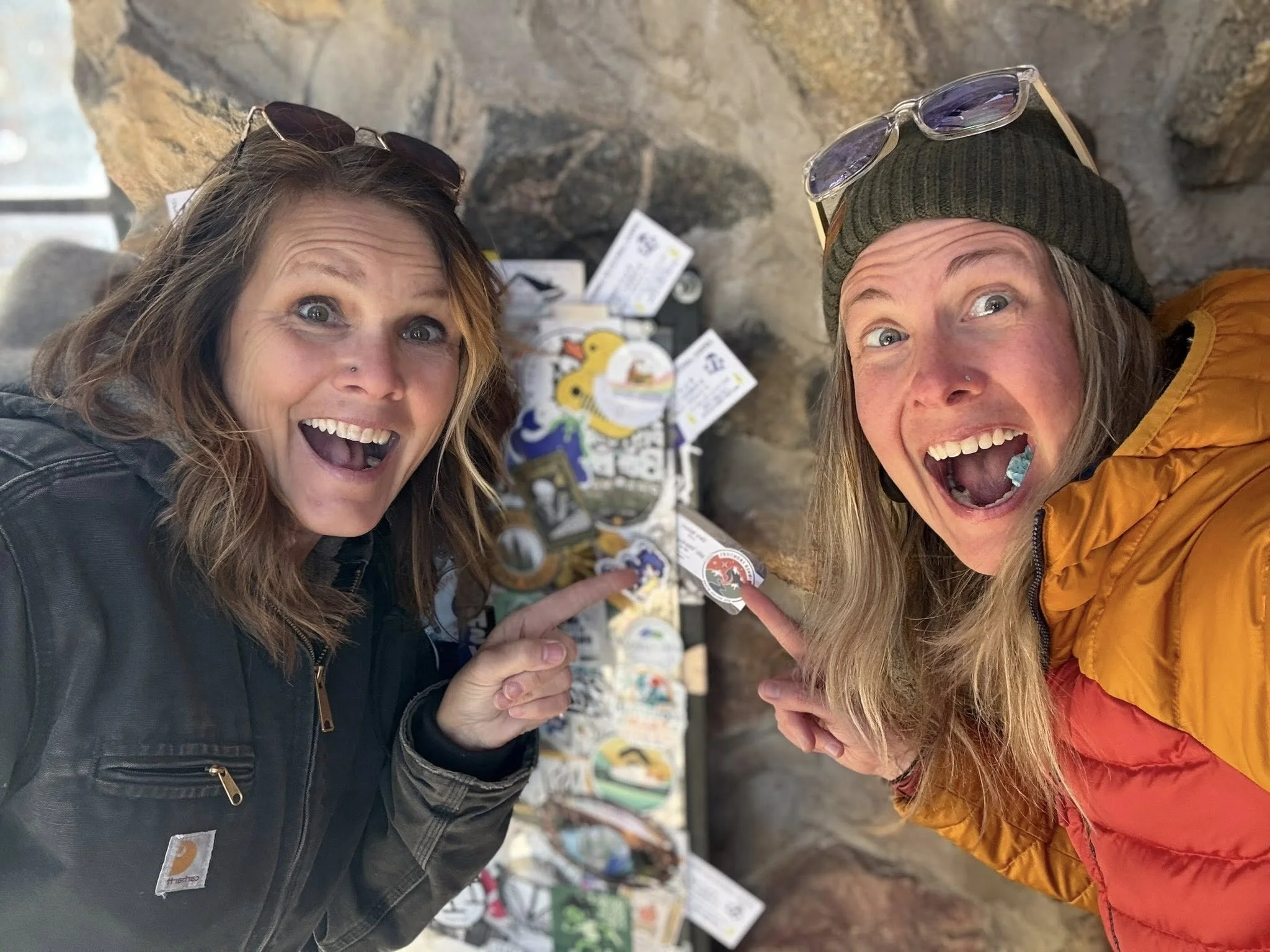 Two women, one with sunglasses on her head and the other wearing glasses on her head, smiling and pointing at a display of various stickers or patches on a wall made of rocks or stone. Both women appear excited or happy.