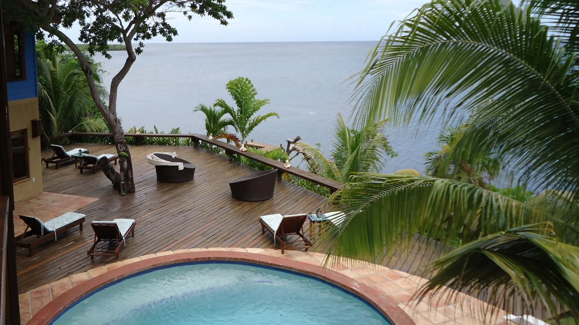 A spacious wooden deck overlooking a body of water, surrounded by tropical plants and trees, with outdoor seating and a hot tub in the foreground.