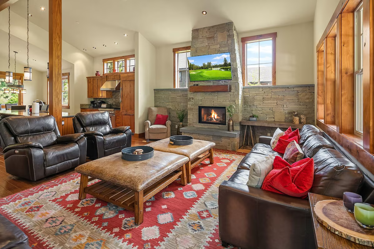 Cozy living room with a leather sofa, armchairs, a stone fireplace, TV, windows, colorful rug, and wooden accents.