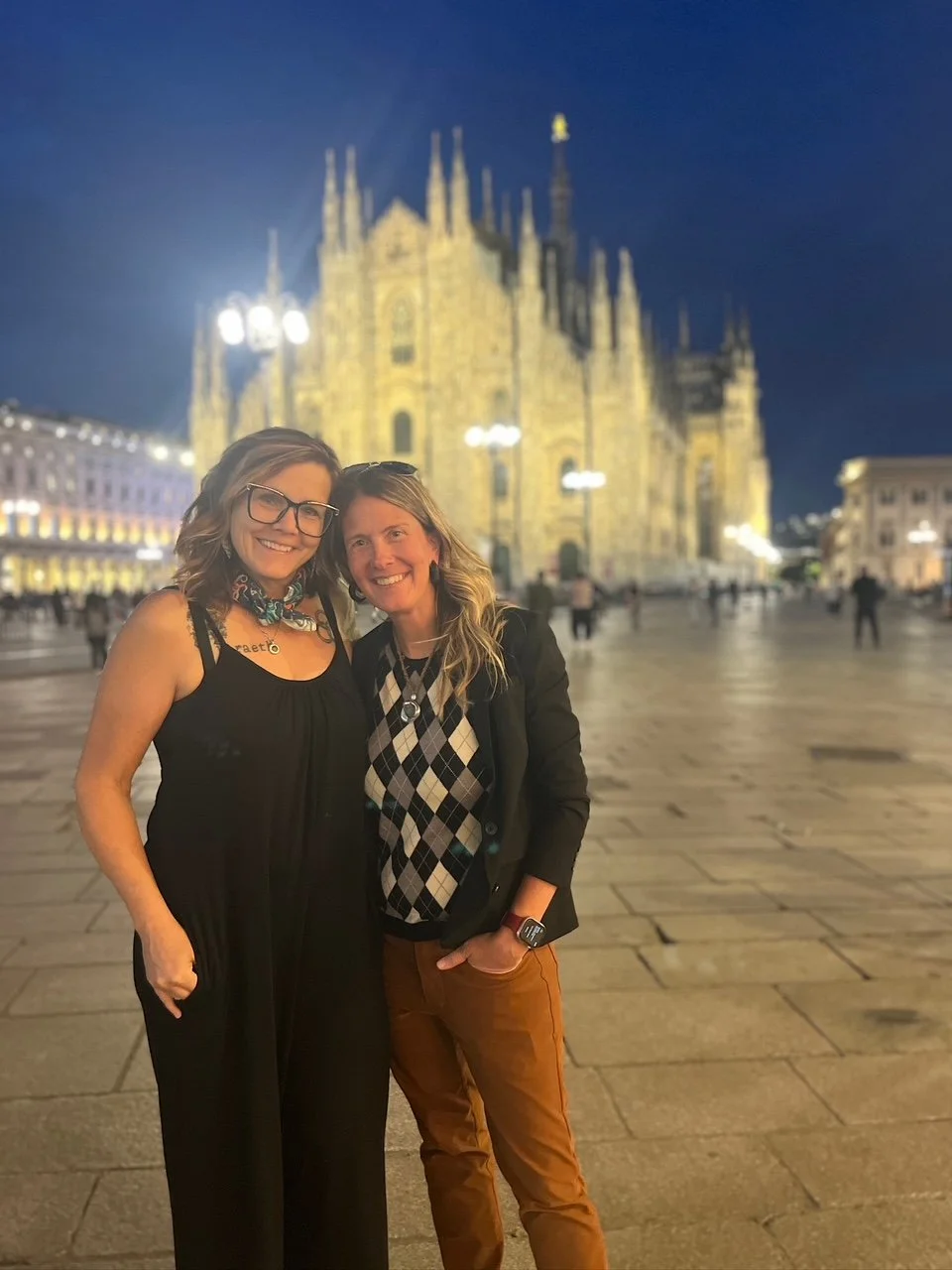 Two women smiling and standing close together in front of the illuminated Milan Cathedral at night, with a clear evening sky and scattered people in the background.
