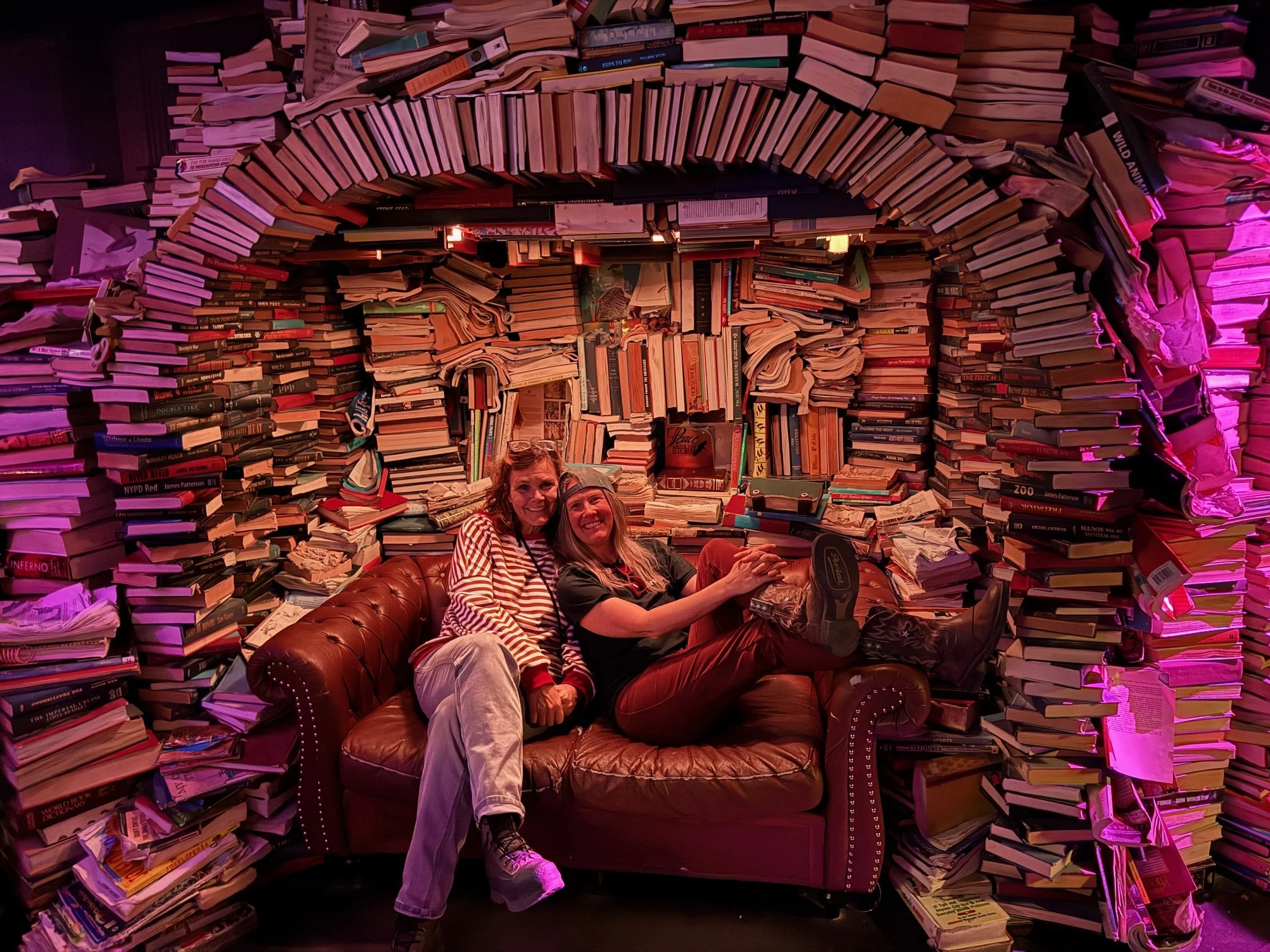 Two women sitting on a brown leather couch in front of a wall covered with stacks of books, illuminated by pink lighting, smiling at the camera.