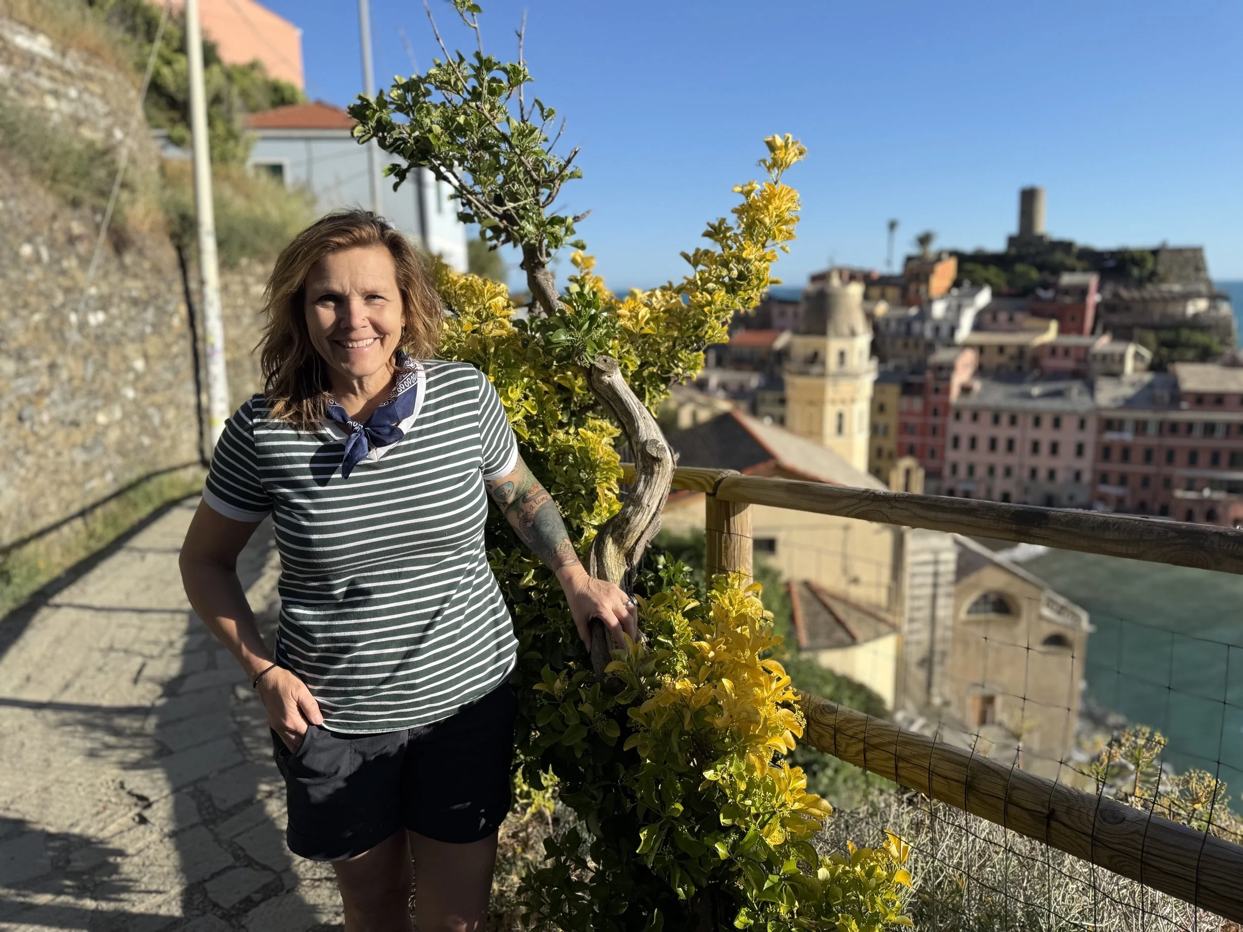 A woman smiling outdoors, standing next to a yellow flowering shrub, with a background of colorful buildings and a blue sky.