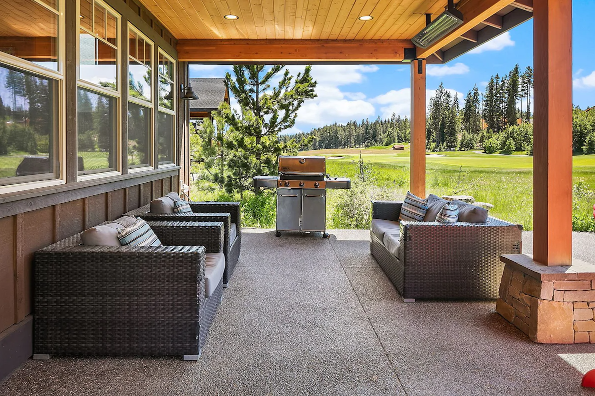 Patio with wicker furniture and a barbecue grill overlooking a green field and trees under a blue sky.