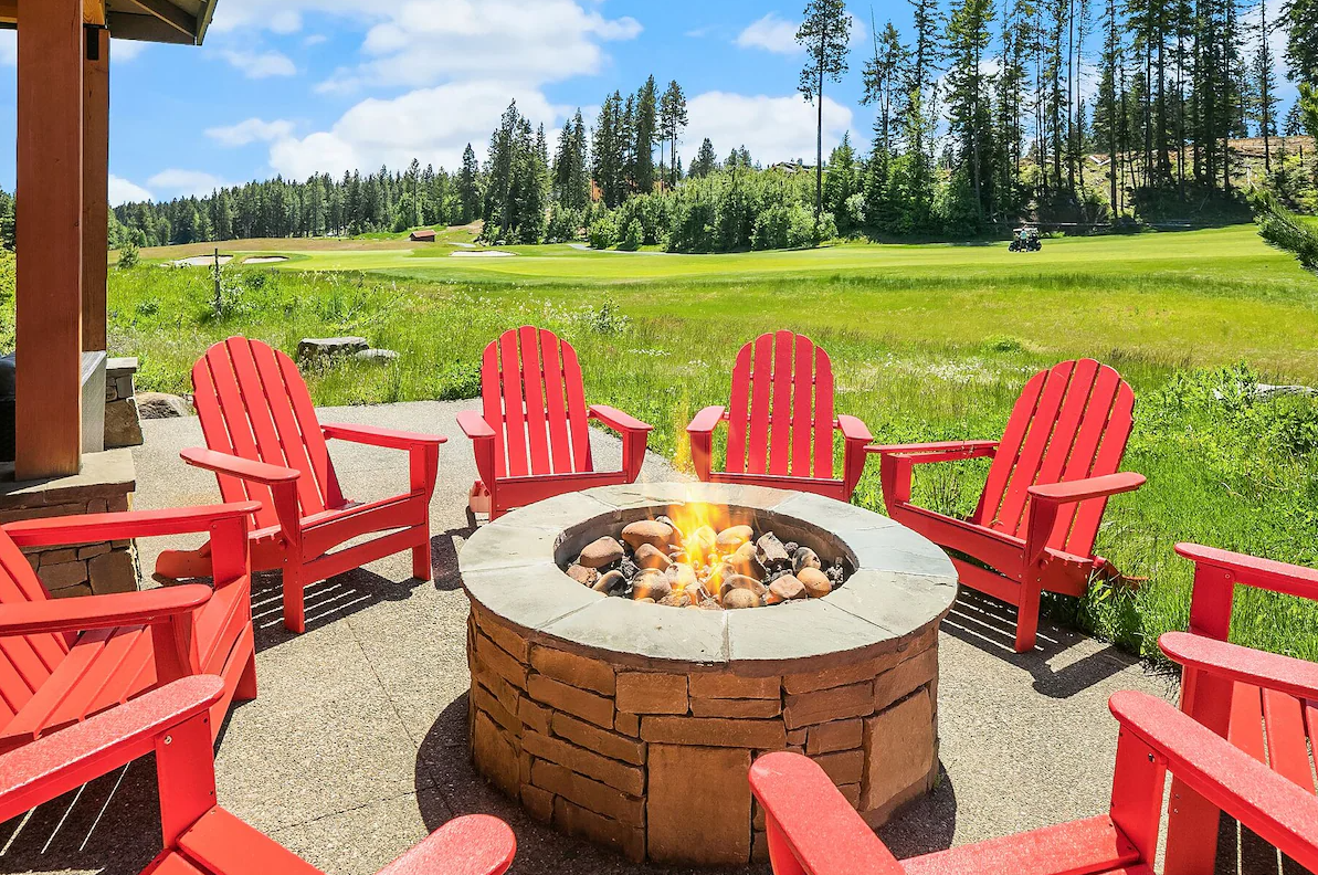 A circle of red Adirondack chairs surrounds a stone fire pit with a small flame, set against a backdrop of a grassy field, trees, and a partly cloudy sky.