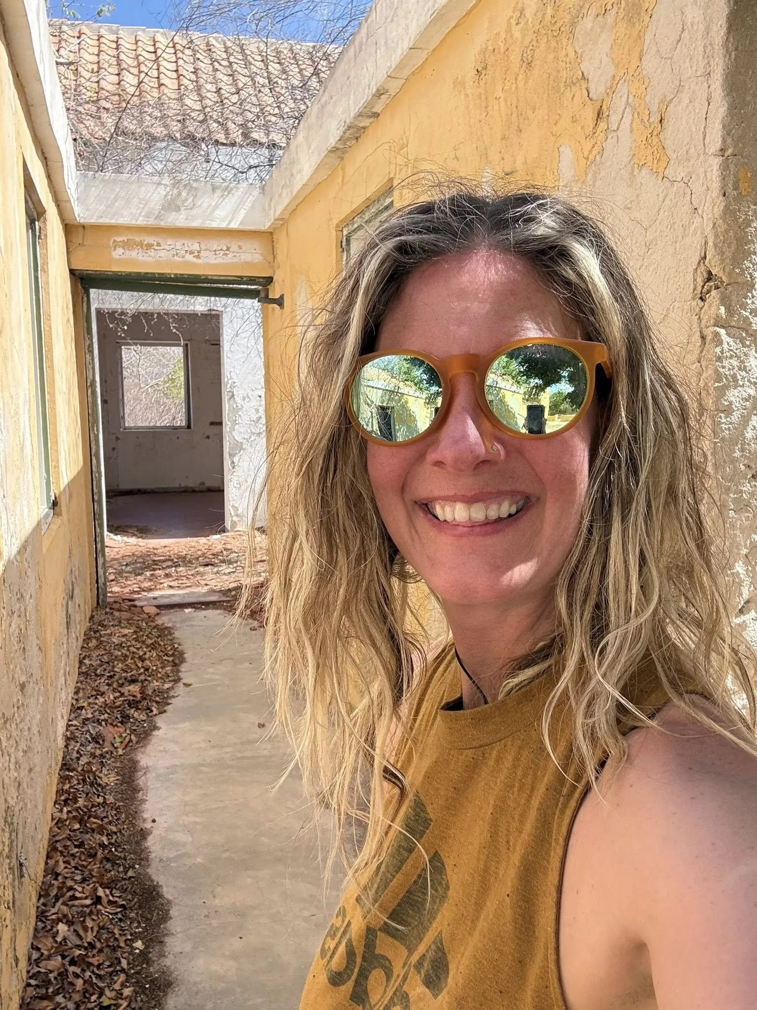 A woman with long curly hair and sunglasses smiling outdoors, standing by a weathered yellow wall with an open doorway behind her.
