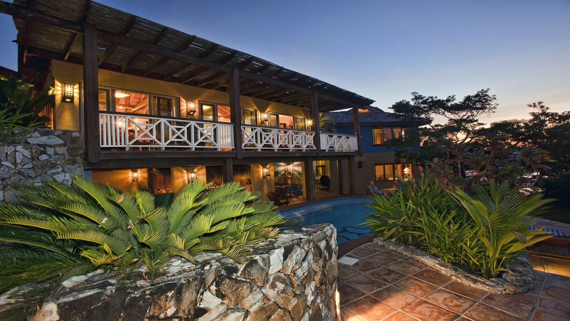 A two-story house with a stone and wood exterior, illuminated by warm lights, surrounded by lush green plants, with a swimming pool in the foreground, during sunset.