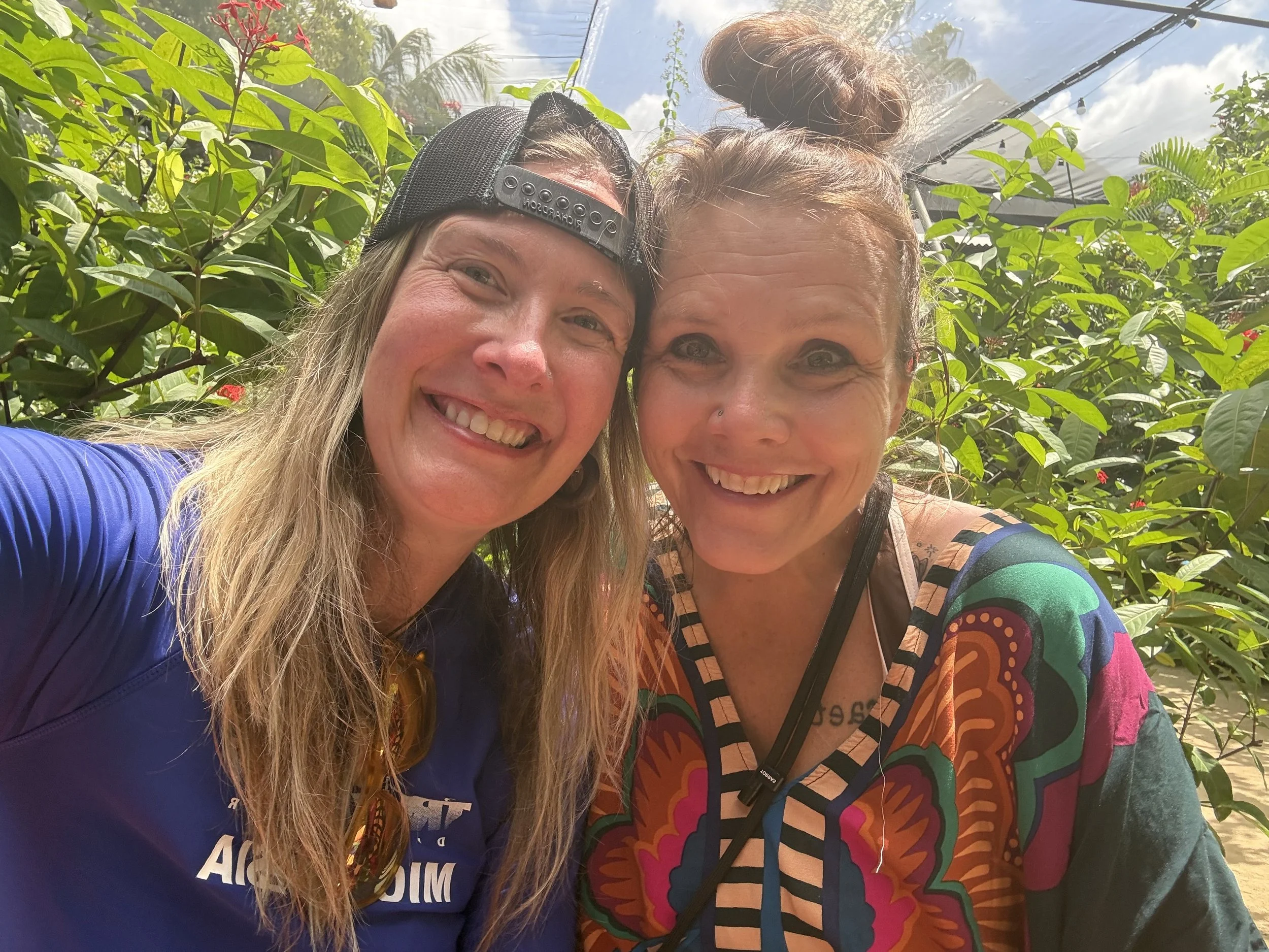 Two smiling women taking a selfie outdoors among green plants under a partly cloudy sky.