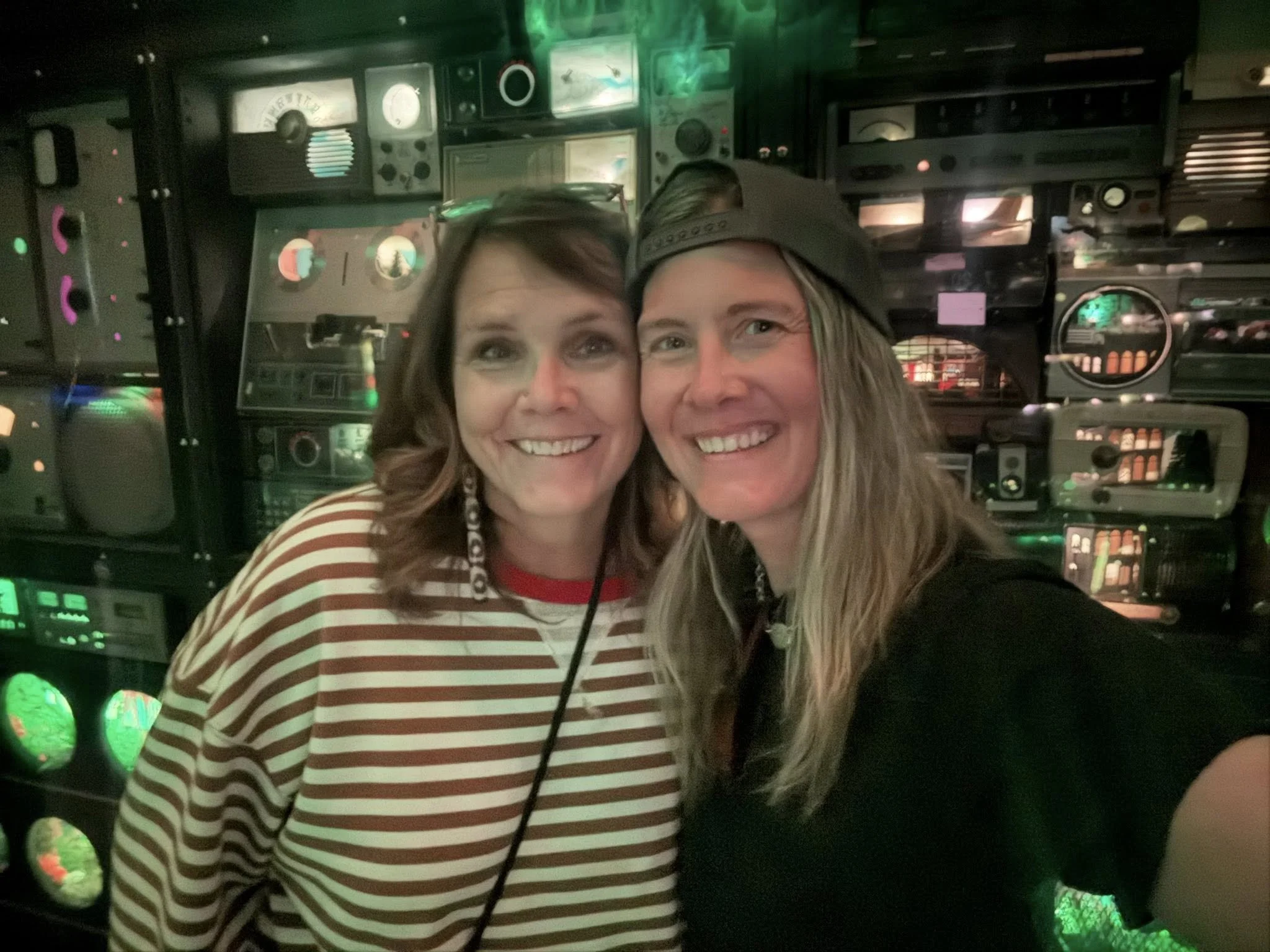 Two women smiling for a selfie in front of a wall of electronic equipment with colorful lights.