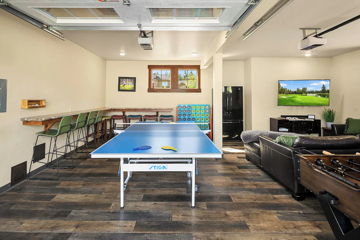 A basement recreation room containing a ping pong table, a dartboard, a bar counter with green chairs, a television, a couch with a green pillow, a foosball table, and a mini fridge, with wood laminate flooring and ceiling-mounted projectors.