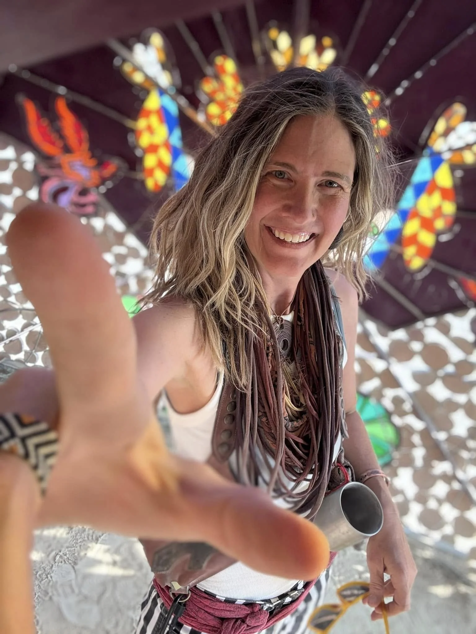 A smiling woman with blonde hair is reaching toward the camera, held in a place that makes it look close. She is wearing a white tank top, striped shorts, and a scarf with red and white patterns. She has a metal cup in her right hand and a yellow lanyard around her neck. In the background, there are colorful stained glass shapes and a dark purple structure.